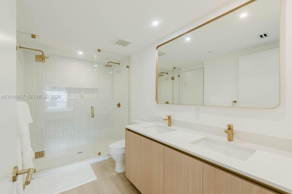 Double vanity with light wood-finish cabinetry, quartz countertop, dual undermount sinks, brass fixtures, and a glass-enclosed shower