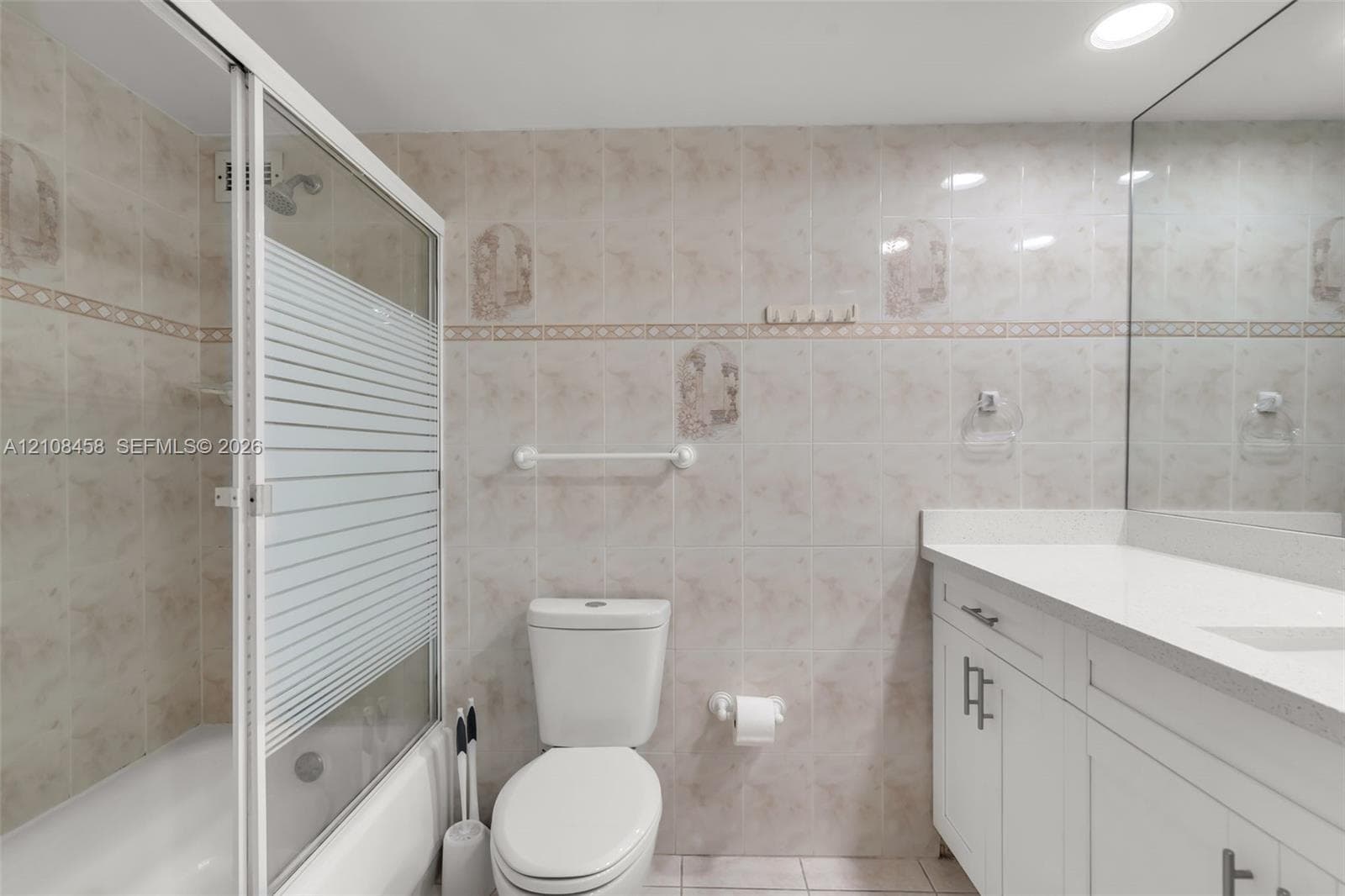 Bathroom featuring white shaker-style cabinetry, quartz countertop with integrated basin, floor-to-ceiling tile walls with decorative accents, and a tub-shower combination with a sliding glass door