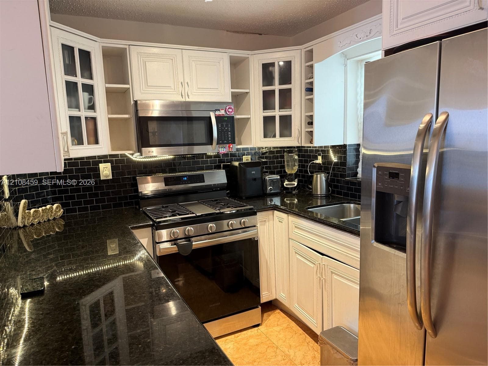 Kitchen featuring white cabinetry with glass-panel display doors, stone countertops, black subway tile backsplash, and stainless steel appliances including a gas range and built-in microwave