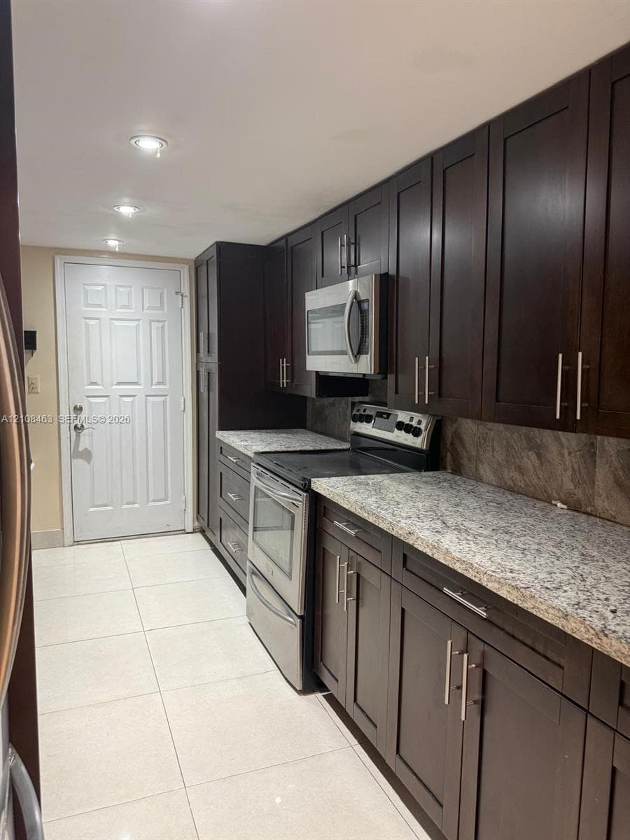 Dark wood-finish cabinetry, stone-finish countertops, stainless steel appliances, tile flooring, and recessed lighting