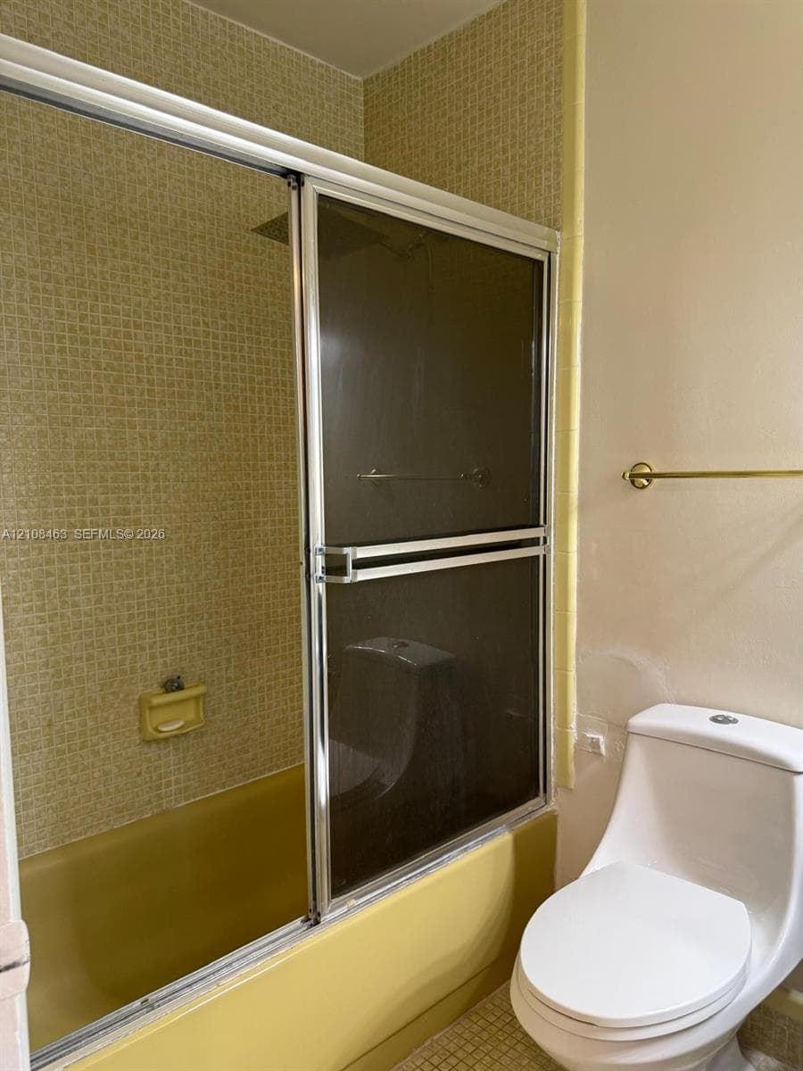 Bathroom featuring a porcelain toilet, a bathtub enclosed by sliding glass doors with metallic trim, grid-patterned wall tile, and an integrated soap dish