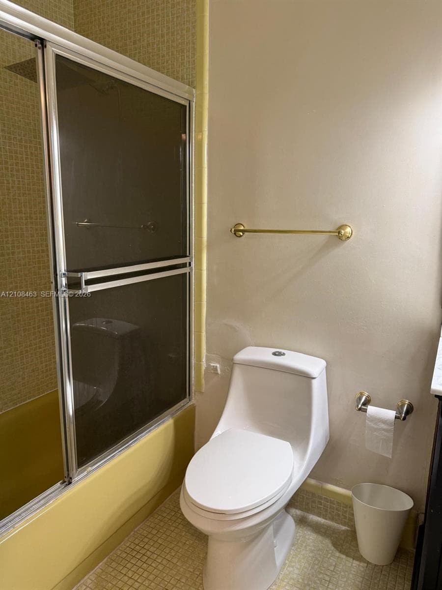 Bathroom featuring a shower-tub combination with sliding glass doors, small-format tile wall cladding, a high-profile white toilet, and gold-finish hardware