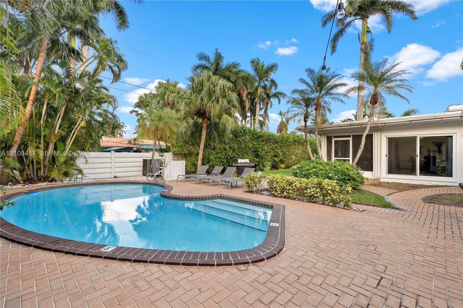 Free-form swimming pool with a dark-tiled border, expansive herringbone-pattern brick paver patio, established palm trees, and perimeter privacy fencing