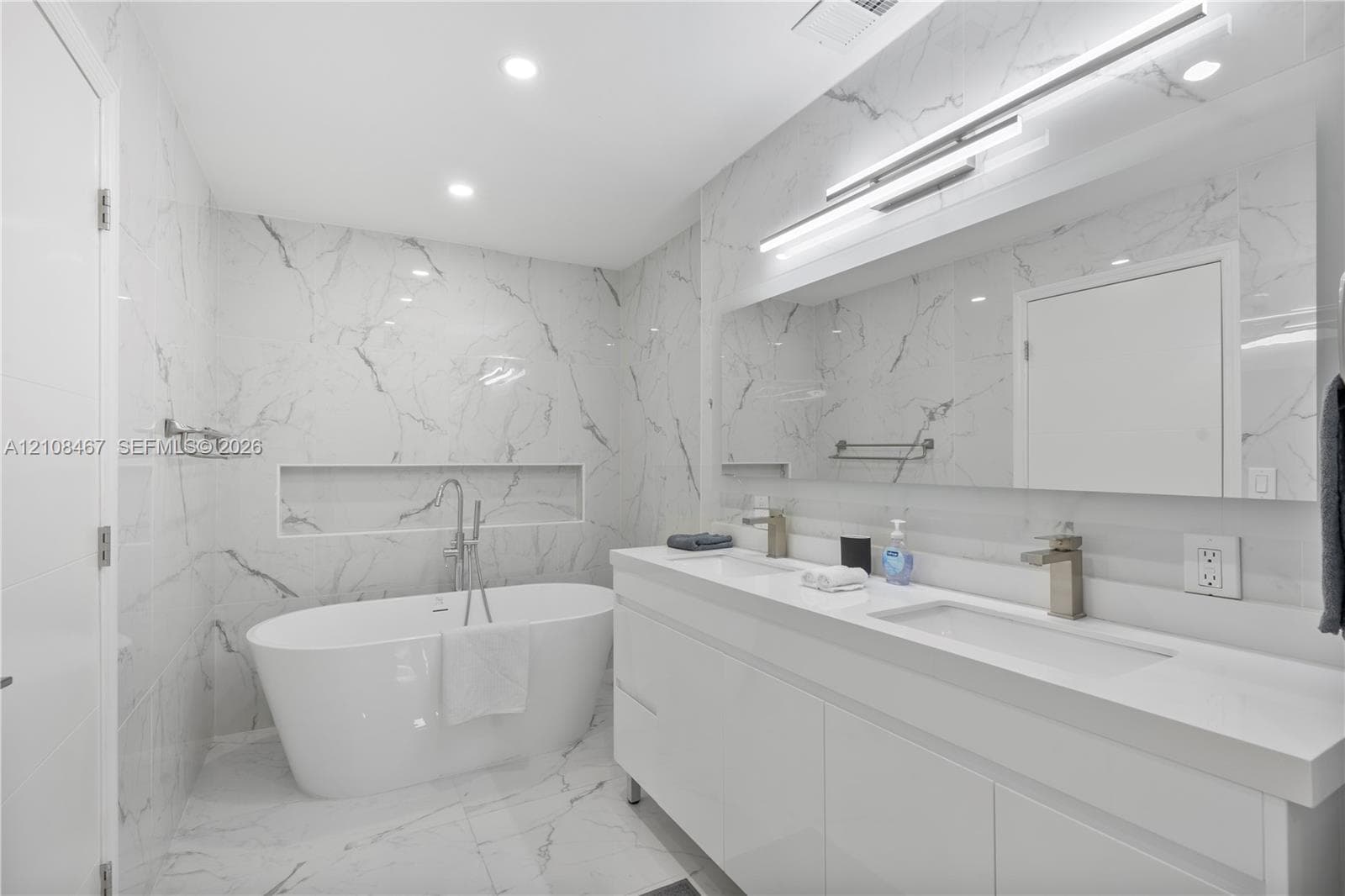 Modern bathroom featuring floor-to-ceiling stone-look tiling, a freestanding soaking tub with a wall-recessed niche, a dual-sink floating vanity, integrated under-cabinet lighting, and a full-width mirror