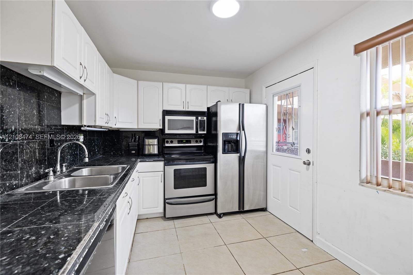Kitchen featuring white shaker-style cabinetry, dark stone-finish countertops, tiled backsplash, stainless steel appliances, and large-format floor tile