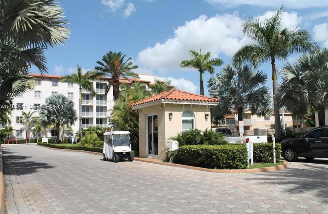 Gated entry featuring a guardhouse with a terracotta-style tile roof, paved driveway, and mature landscaping