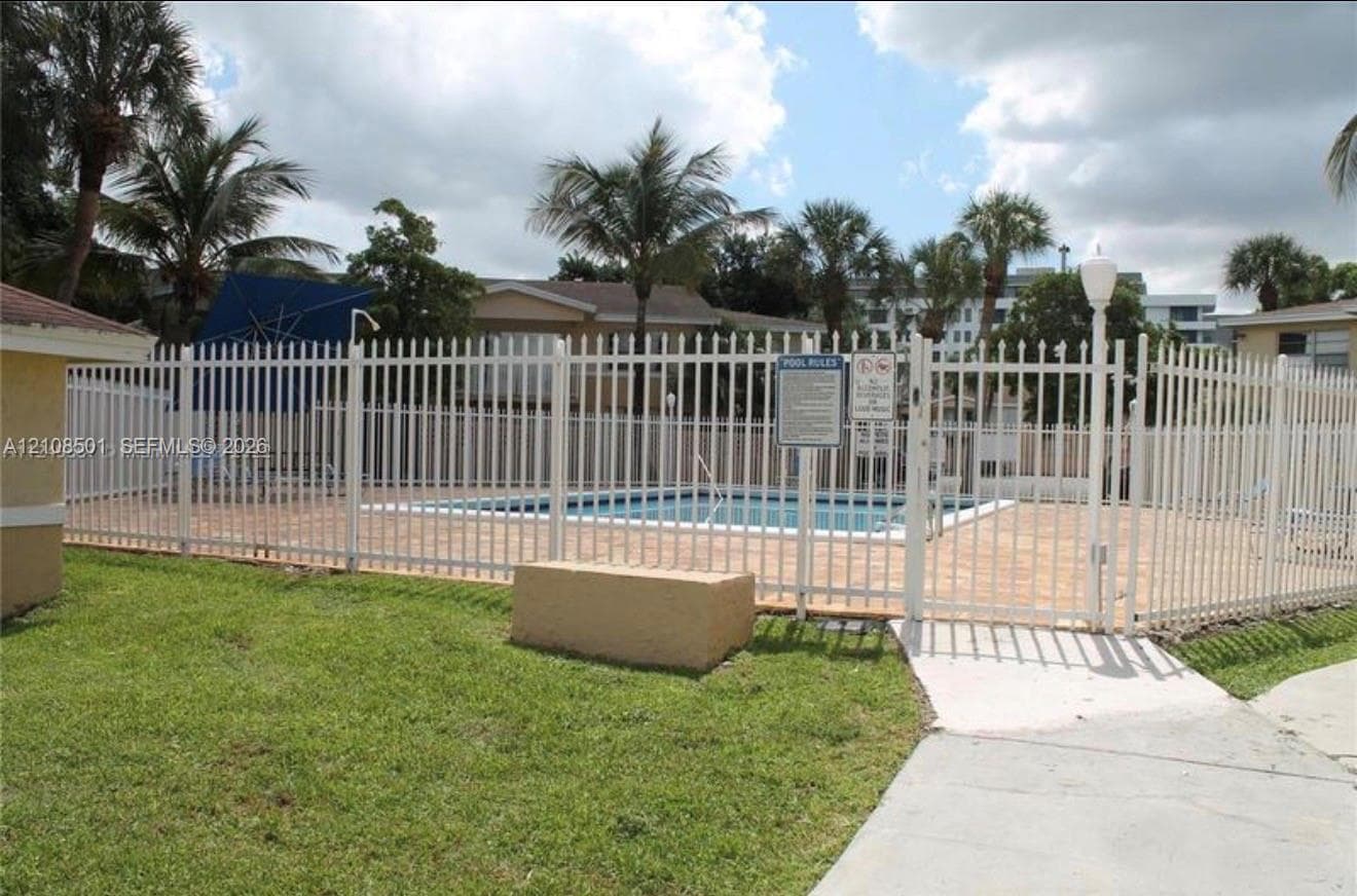 Outdoor swimming pool enclosed by metal fencing, featuring a concrete pool deck, manicured lawn, and perimeter landscaping with palm trees