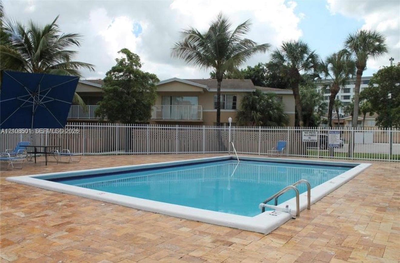In-ground swimming pool featuring a white coping border, stainless steel pool ladder, and surrounding stone paver deck enclosed by white metal fencing