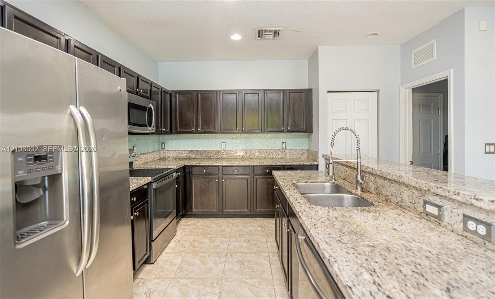 Kitchen featuring dark wood cabinetry, stone-finish countertops, stainless steel appliances, tiled backsplash, and a double-basin undermount sink with a pull-down faucet