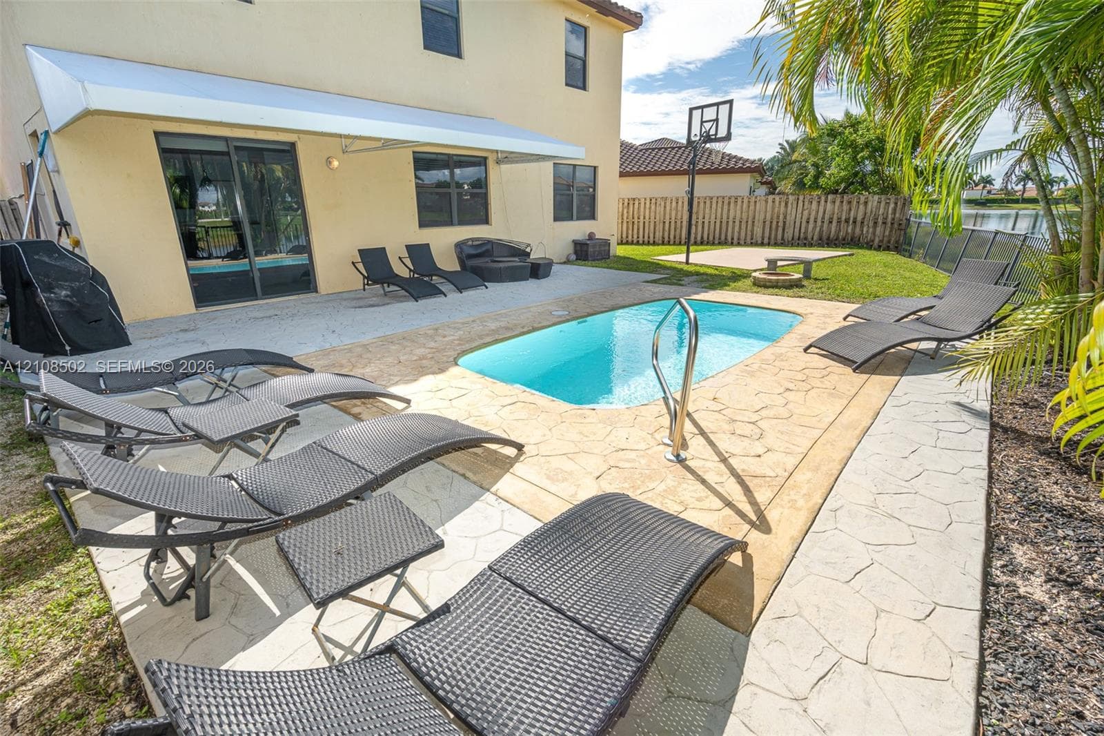 Backyard featuring a concrete pool with a metal handrail, a stamped concrete patio, a retractable awning, and a built-in basketball hoop