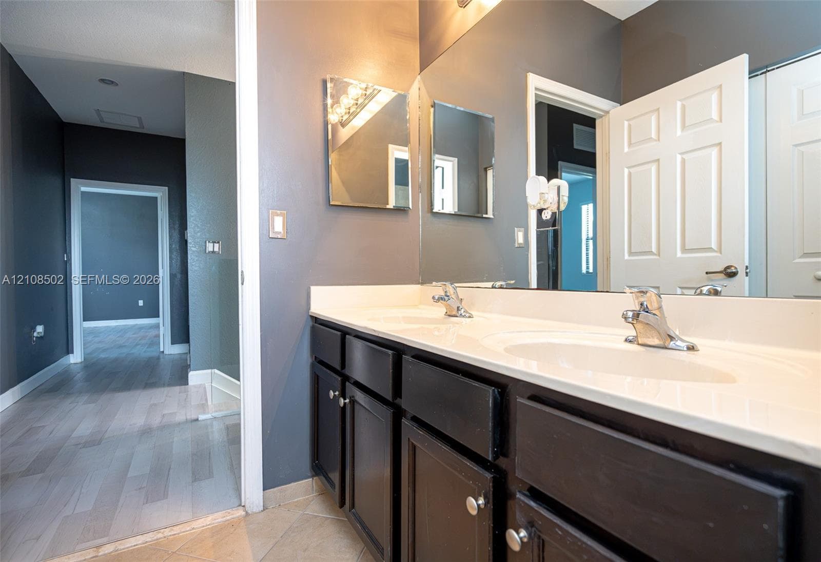 Bathroom vanity featuring dark-toned cabinetry, dual undermount sinks, a solid-surface countertop, full-width wall mirror, and chrome-finish hardware