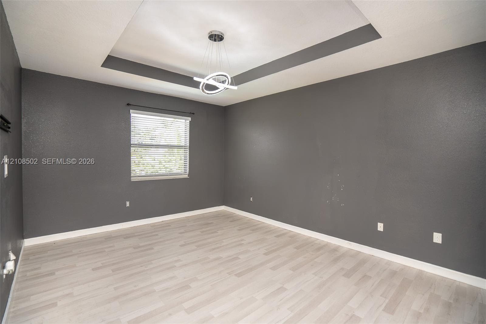 Tray ceiling featuring a contemporary circular chandelier, light-toned wood-finish flooring, and single-pane window with installed blinds