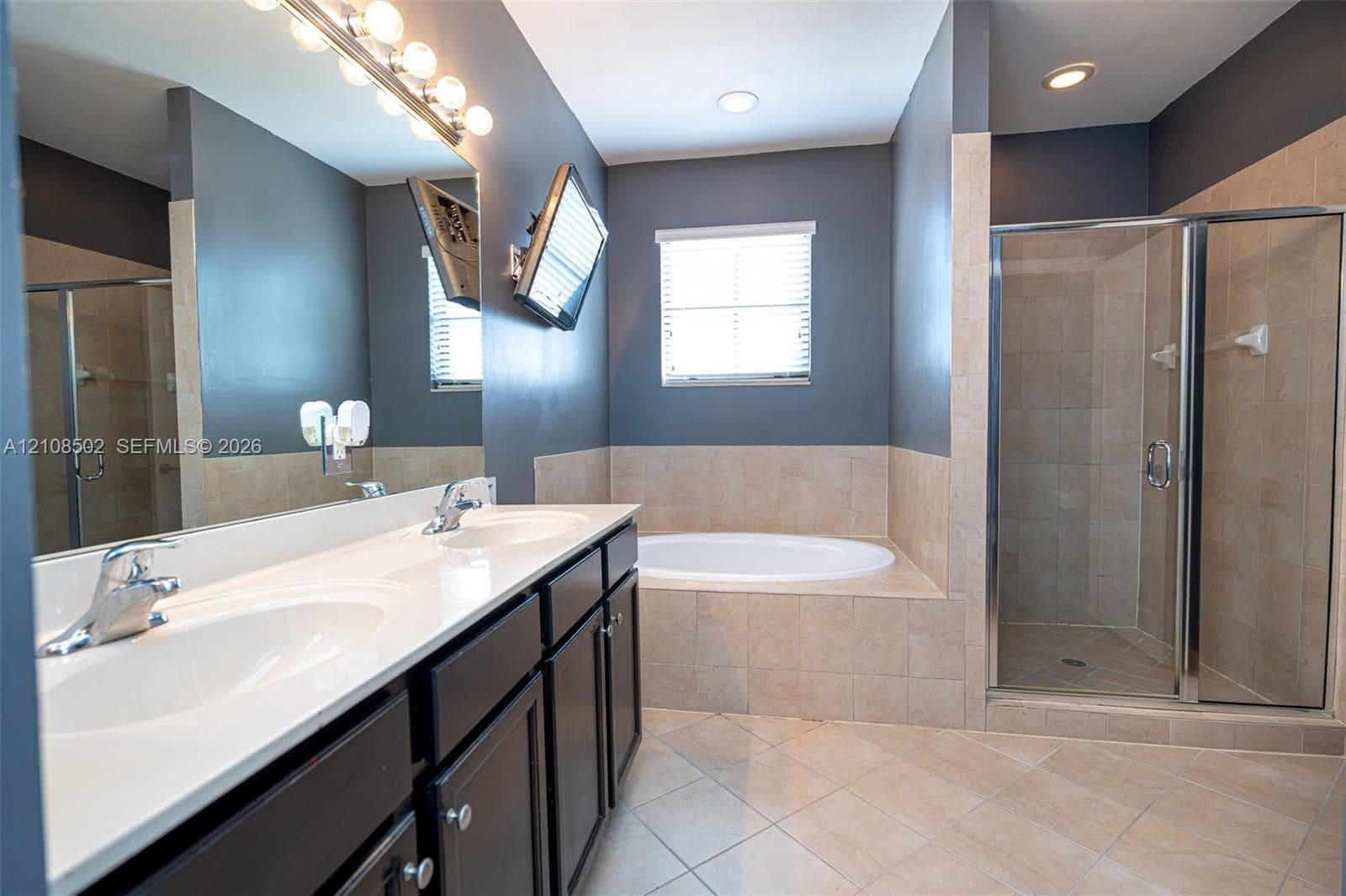 Bathroom featuring a dual-sink vanity with light-toned countertops, a deep soaking tub with tile surround, and a glass-enclosed walk-in shower