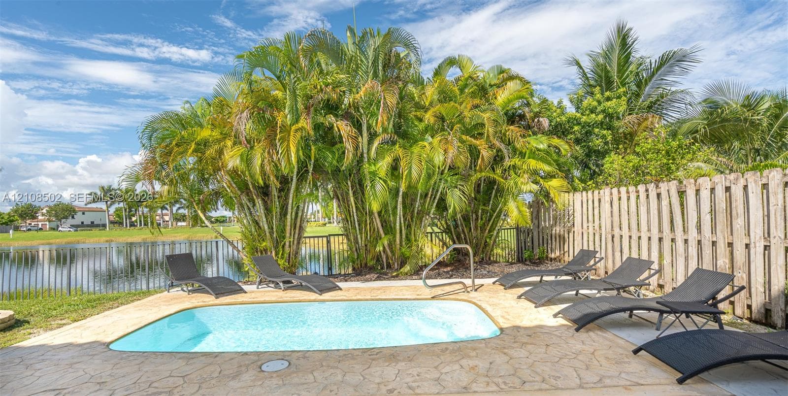 In-ground swimming pool with stone-finish decking, black metal perimeter fencing, and mature tropical landscaping