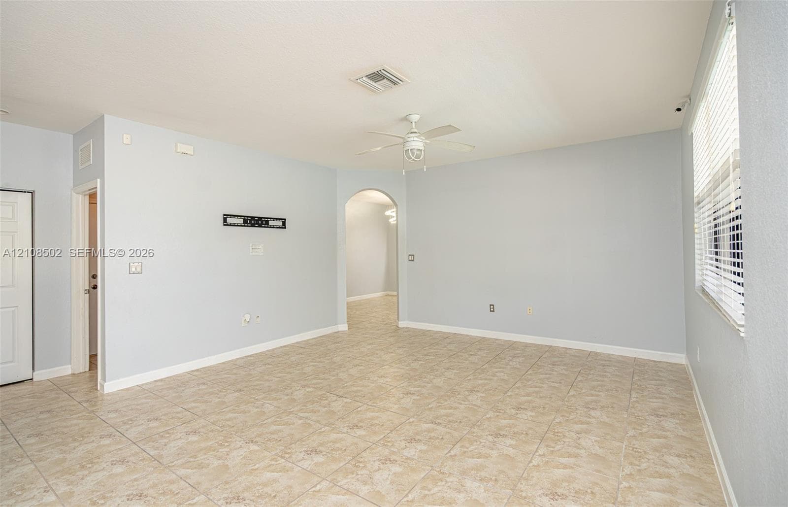 Neutral-toned interior space featuring light-colored tile flooring, an arched doorway, a ceiling fan with integrated lighting, and horizontal window blinds