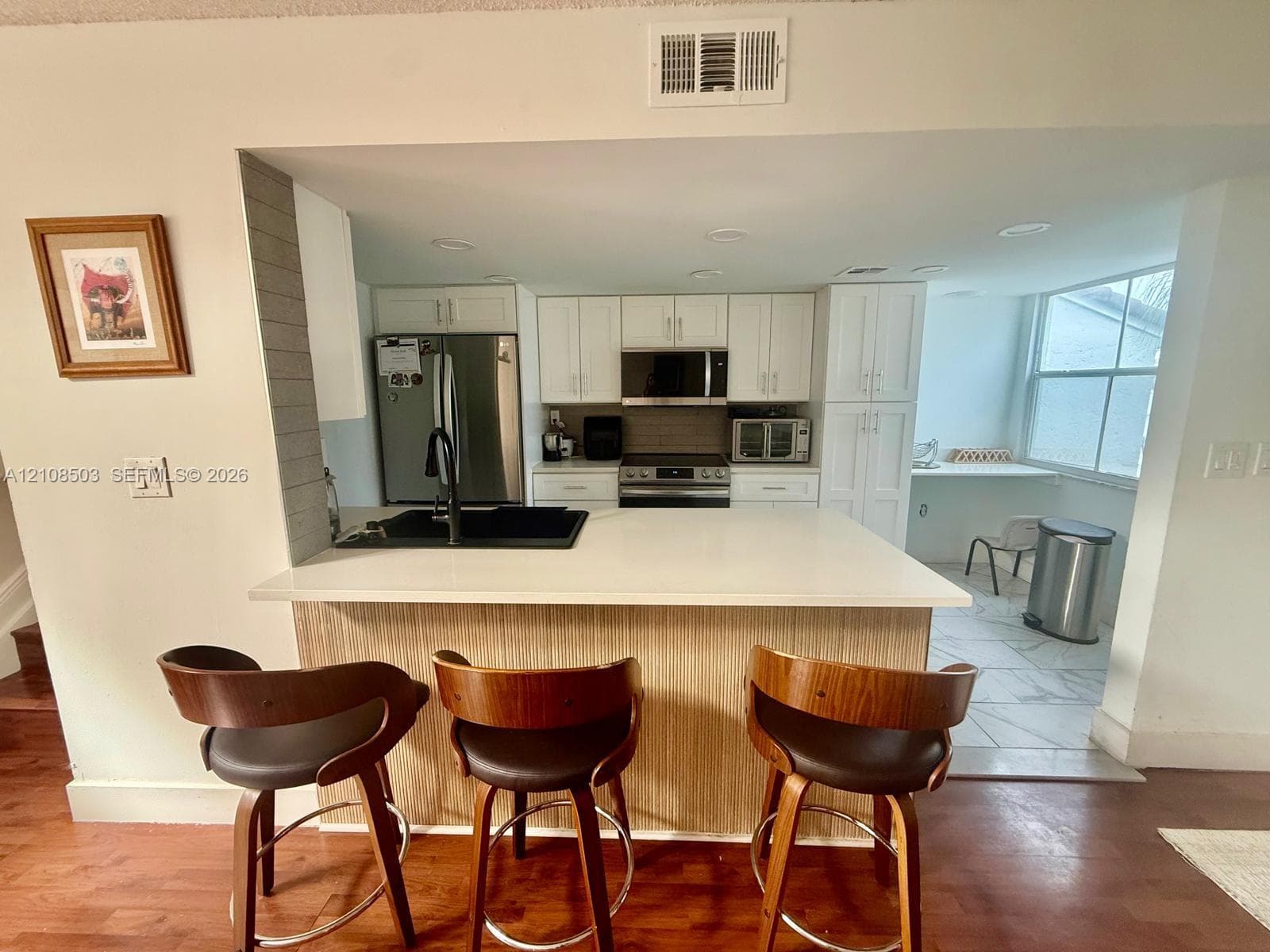 Kitchen featuring white cabinetry, solid-surface countertops, integrated stainless steel appliances, a black undermount sink with a matte black faucet, and wood-finish cabinetry detailing