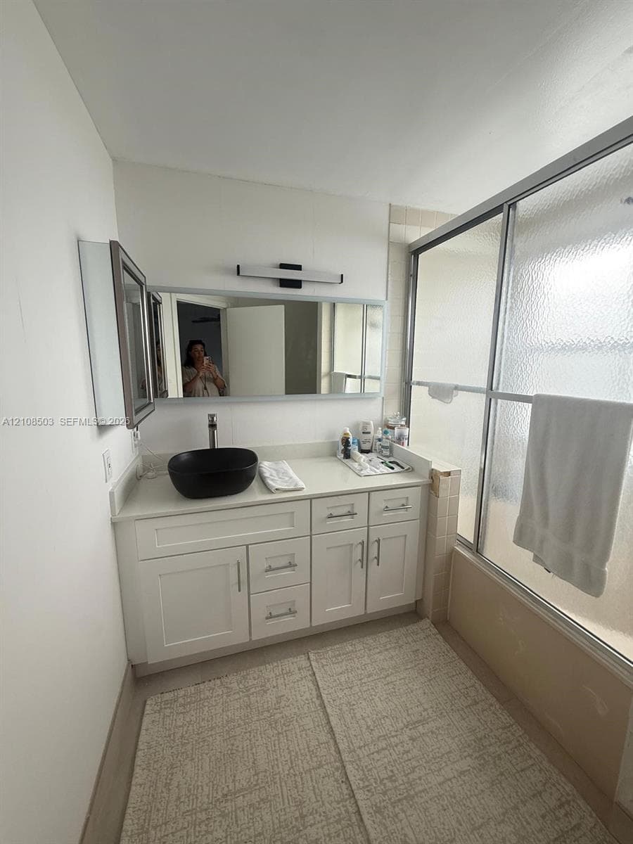 White cabinetry vanity featuring a solid surface countertop, matte black vessel sink, wall-mounted rectangular mirror, and a sliding glass shower door