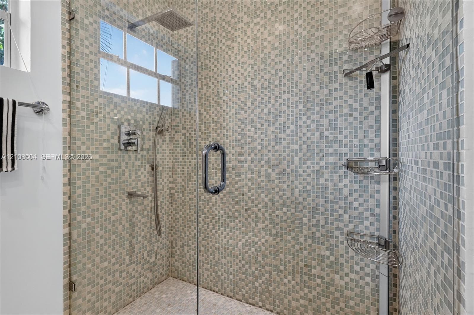 Walk-in shower featuring a frameless glass door, multi-tone mosaic wall tile, overhead rain shower head, handheld shower attachment, and integrated corner shelving