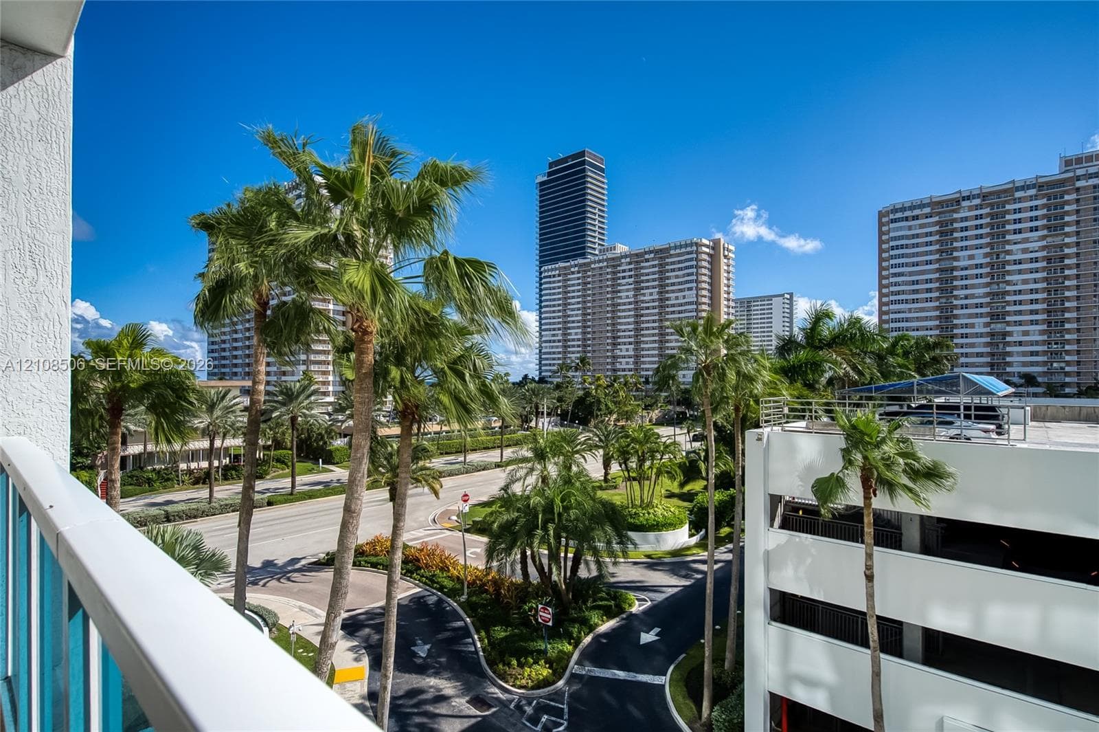 Exterior balcony with white metal railings, overlooking an urban streetscape featuring mature palm trees and high-rise architecture