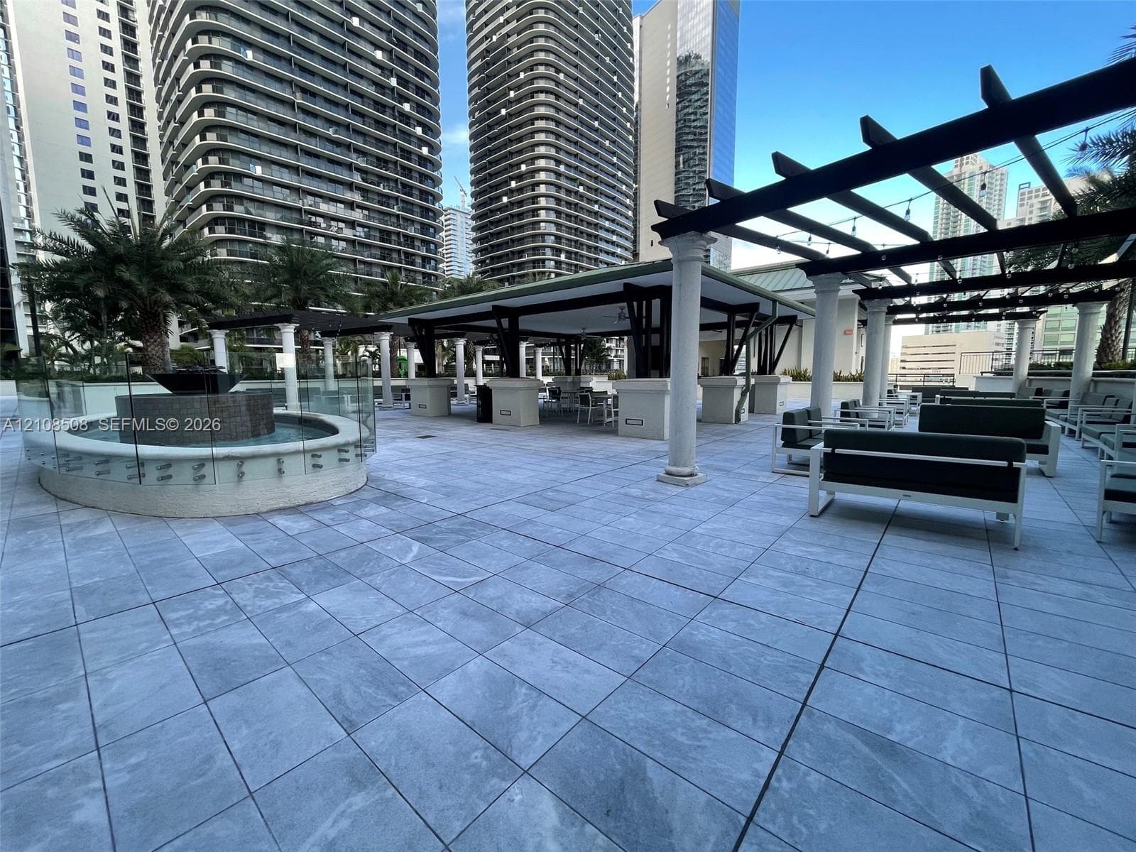 Outdoor terrace featuring large-format stone-look tiling, a circular stone-clad water feature with glass surround, and a covered pergola supported by classic columns