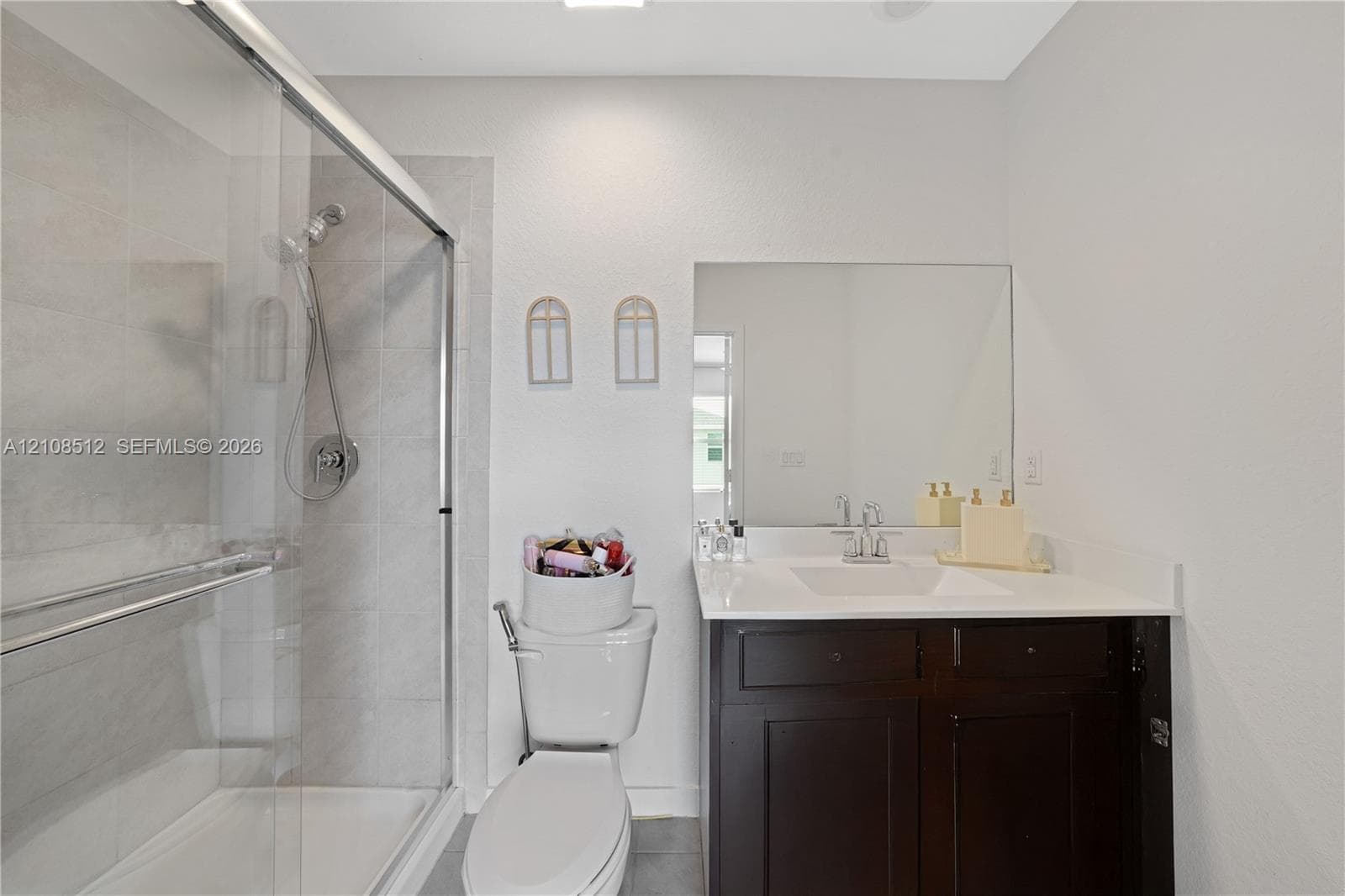 Walk-in shower with sliding glass enclosure and tiled walls, dark-toned vanity cabinet, white solid-surface countertop with integrated basin, and expansive frameless wall mirror