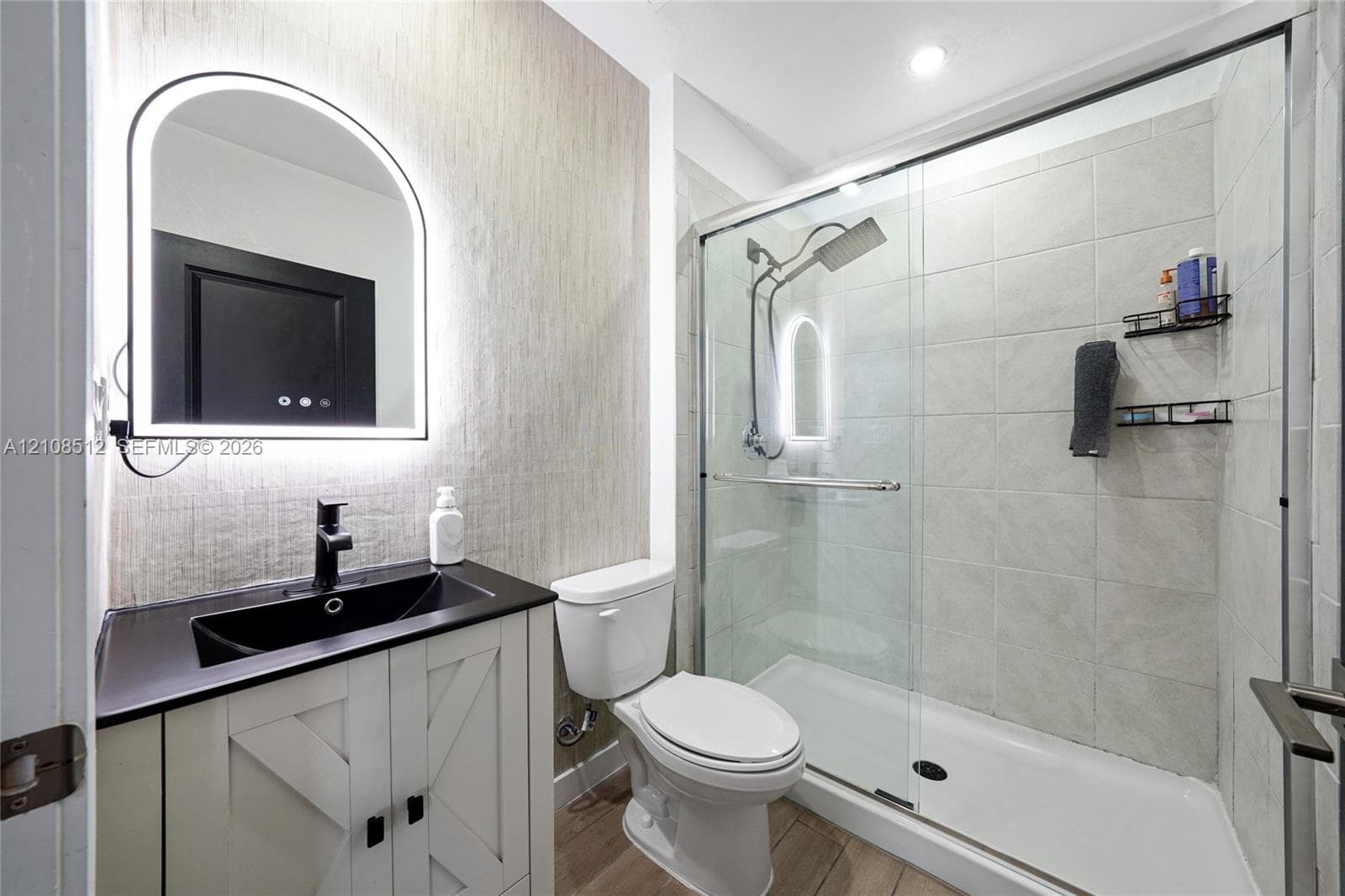 Bathroom featuring a vanity with a dark countertop and integrated sink, arch-top lighted mirror, wood-finish flooring, and a walk-in shower with a glass enclosure and tiled walls