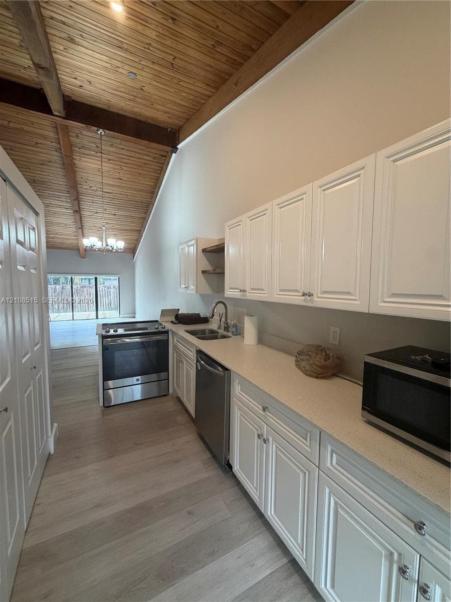 Vaulted ceiling with exposed wood beams, white cabinetry, solid-surface countertops, wood-finish flooring, and stainless steel appliances
