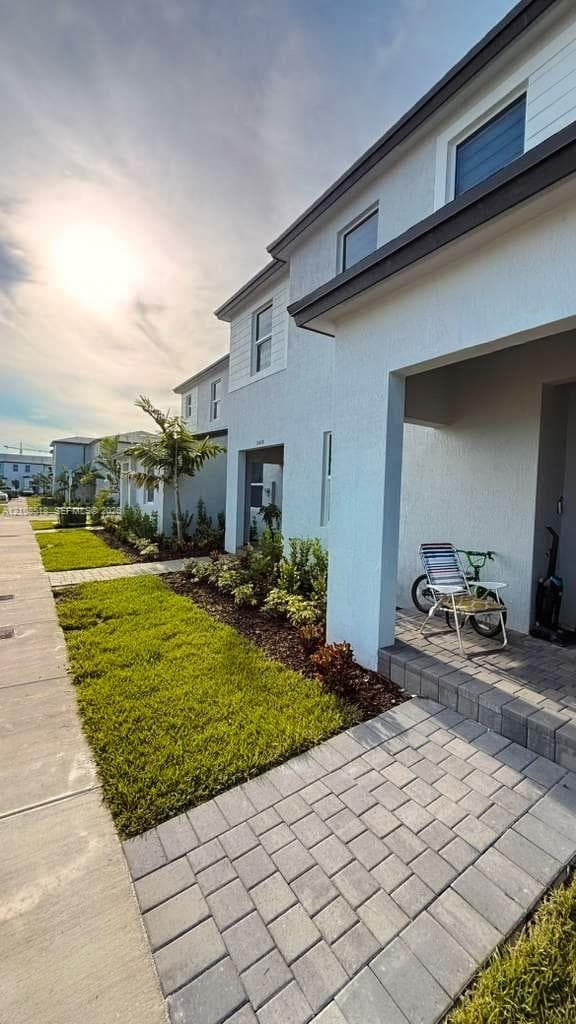 Exterior facade featuring light-toned stucco and horizontal siding, covered entryway with paver flooring, and landscaped front yard with turf and mulch beds