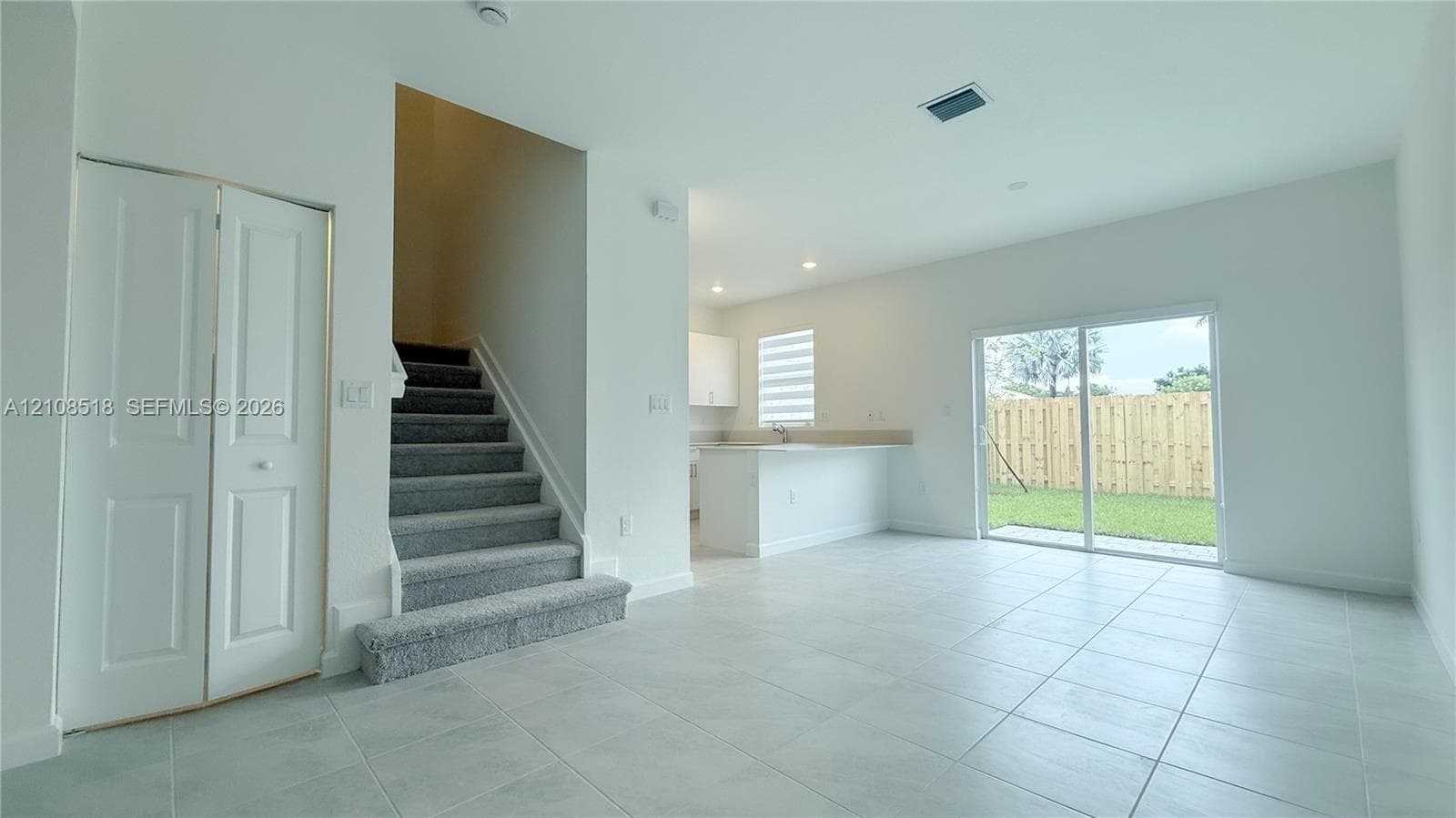Open-concept interior featuring neutral tile flooring, carpeted staircase with white railings, sliding glass doors, built-in storage closet, and kitchen peninsula