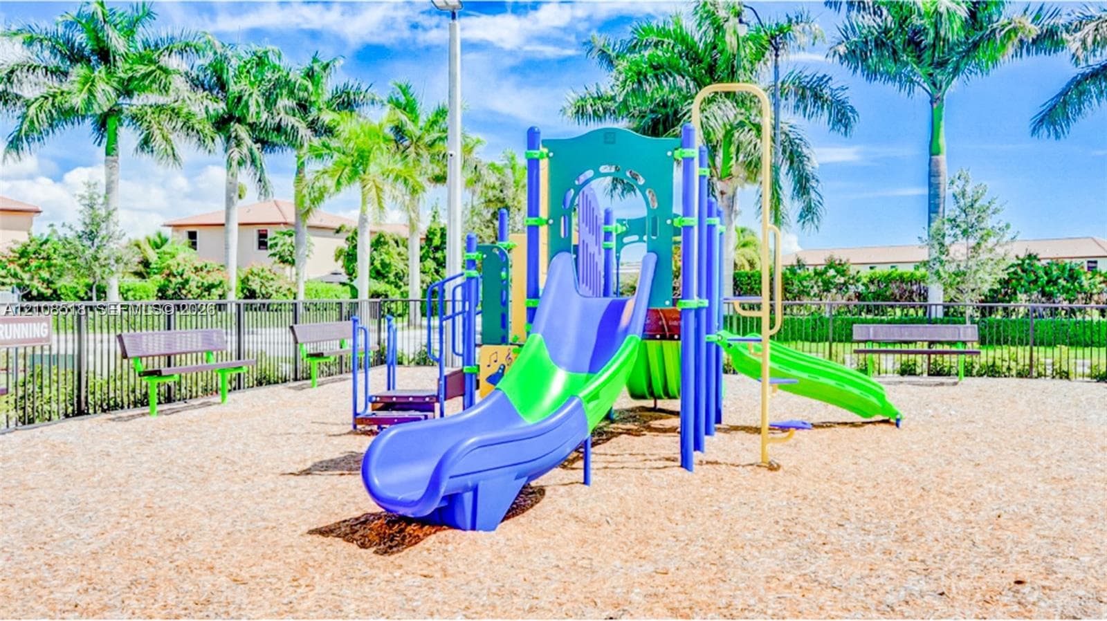 Community playground featuring a multi-level structure with slides, integrated benches, and palm-lined landscaping, set within a fenced area with wood chip ground cover