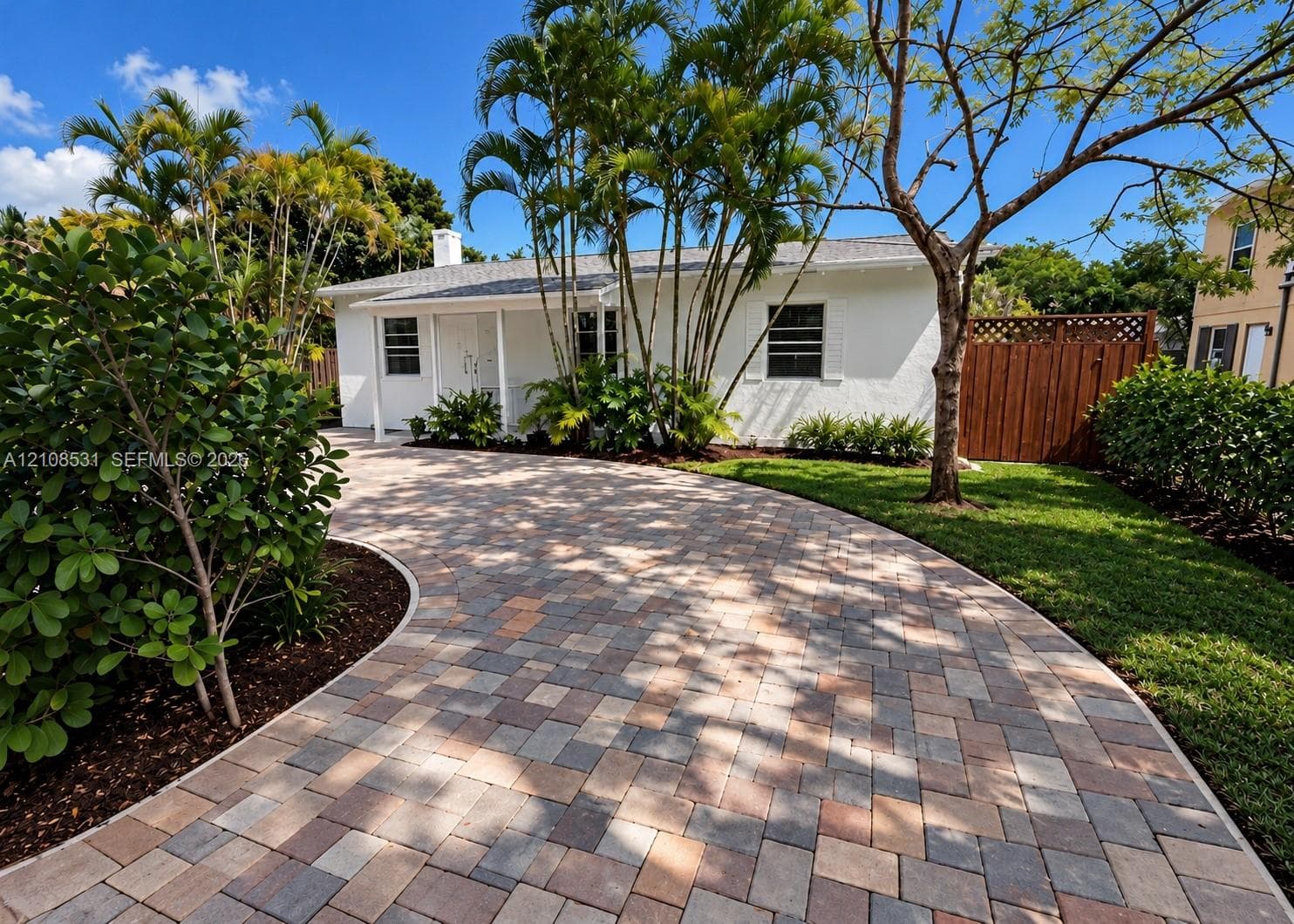 Single-story exterior featuring a multi-tone paver driveway, white stucco finish, front covered porch, and mature palm landscaping