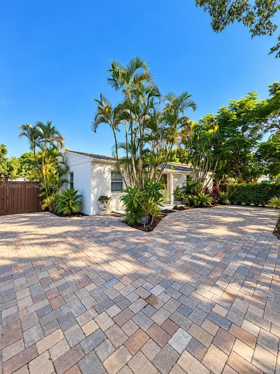 Exterior facade featuring white painted siding, expansive paver driveway, mature palm trees, and perimeter privacy fencing