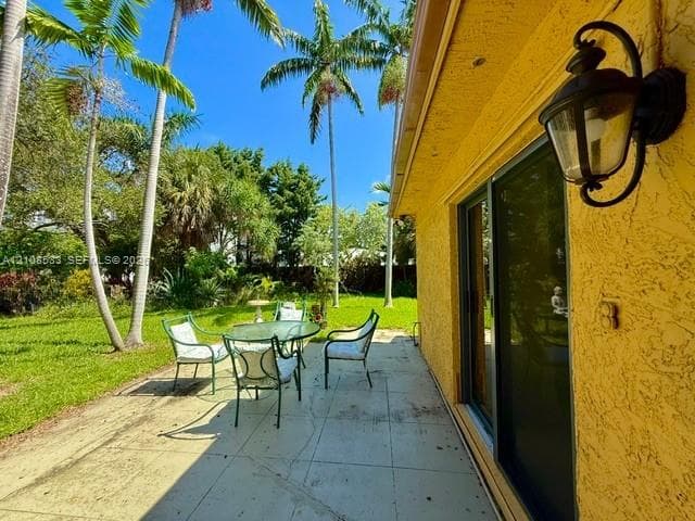 Exterior patio featuring a concrete surface, stucco siding, sliding glass doors, and a wall-mounted coach light, overlooking a lawn with mature palm trees