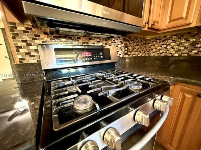 Stainless steel gas range with a built-in microwave, dark stone countertops, mosaic tile backsplash, and wood-finish cabinetry
