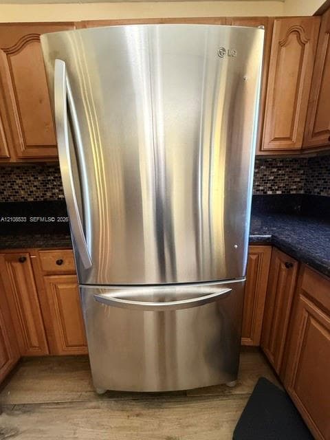 Stainless steel bottom-freezer refrigerator, dark-toned stone countertops, multi-colored mosaic tile backsplash, and wood-finish cabinetry