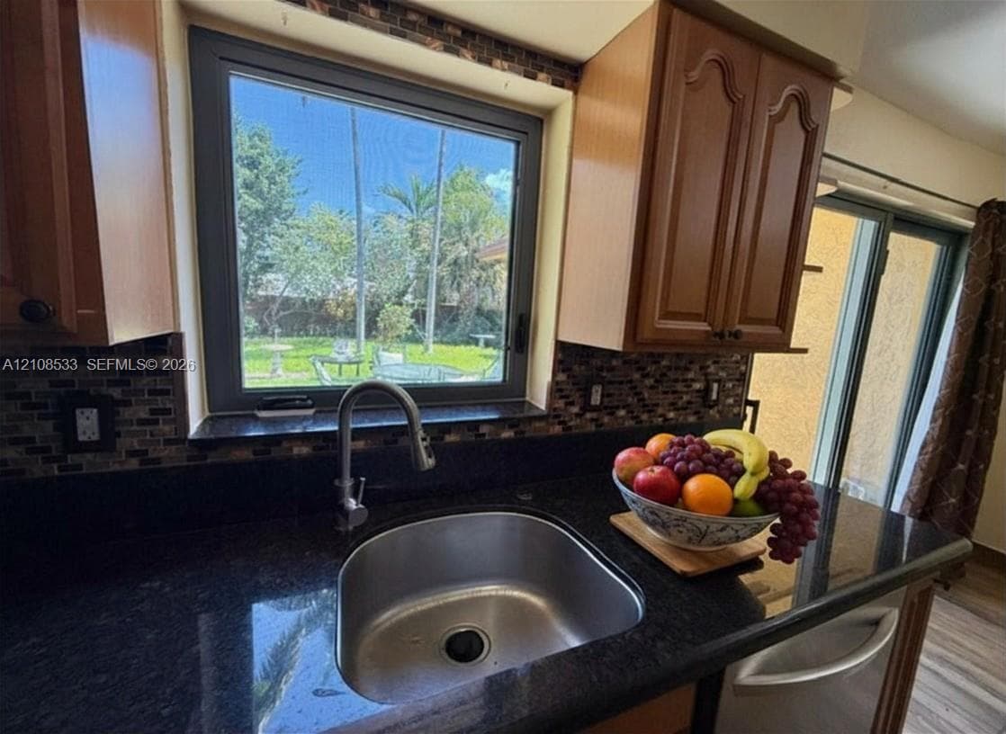 Kitchen featuring dark stone countertops, mosaic tile backsplash, wood-finish cabinetry, stainless steel undermount sink with high-arc faucet, and oversized window