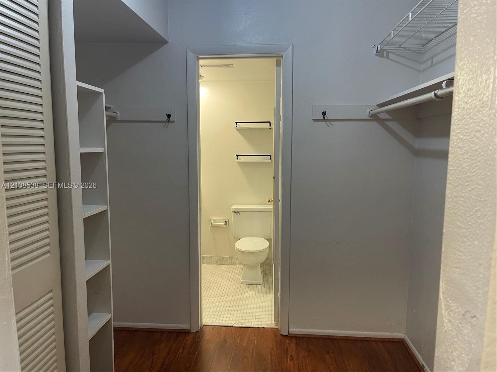 Walk-in closet featuring wood-finish flooring, built-in shelving, and dual hanging rods, with access to a bathroom finished with mosaic tile flooring and wall-mounted shelving