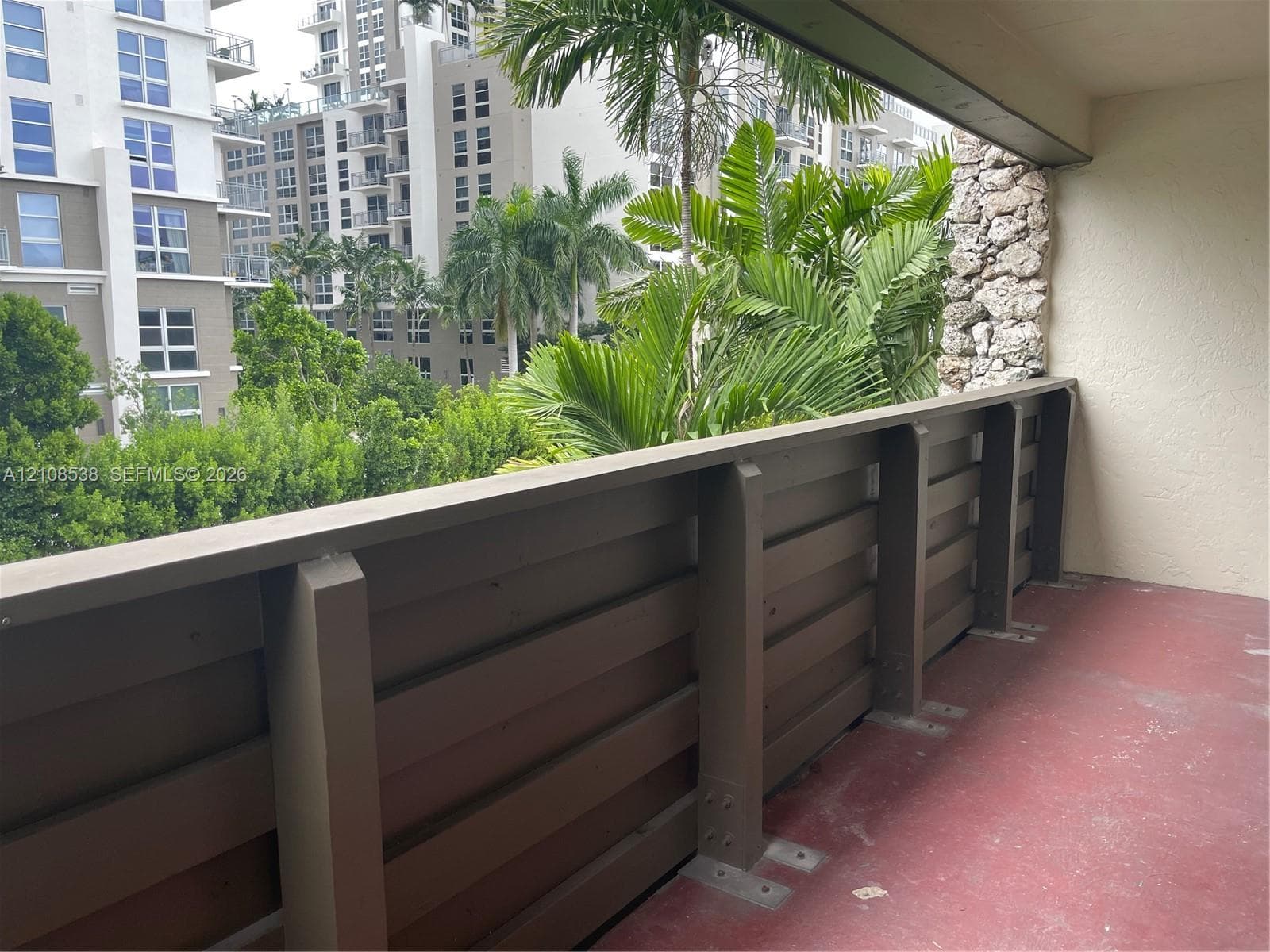 Covered balcony featuring horizontal wood-finish railing, textured stone accent wall, and palm tree scenery