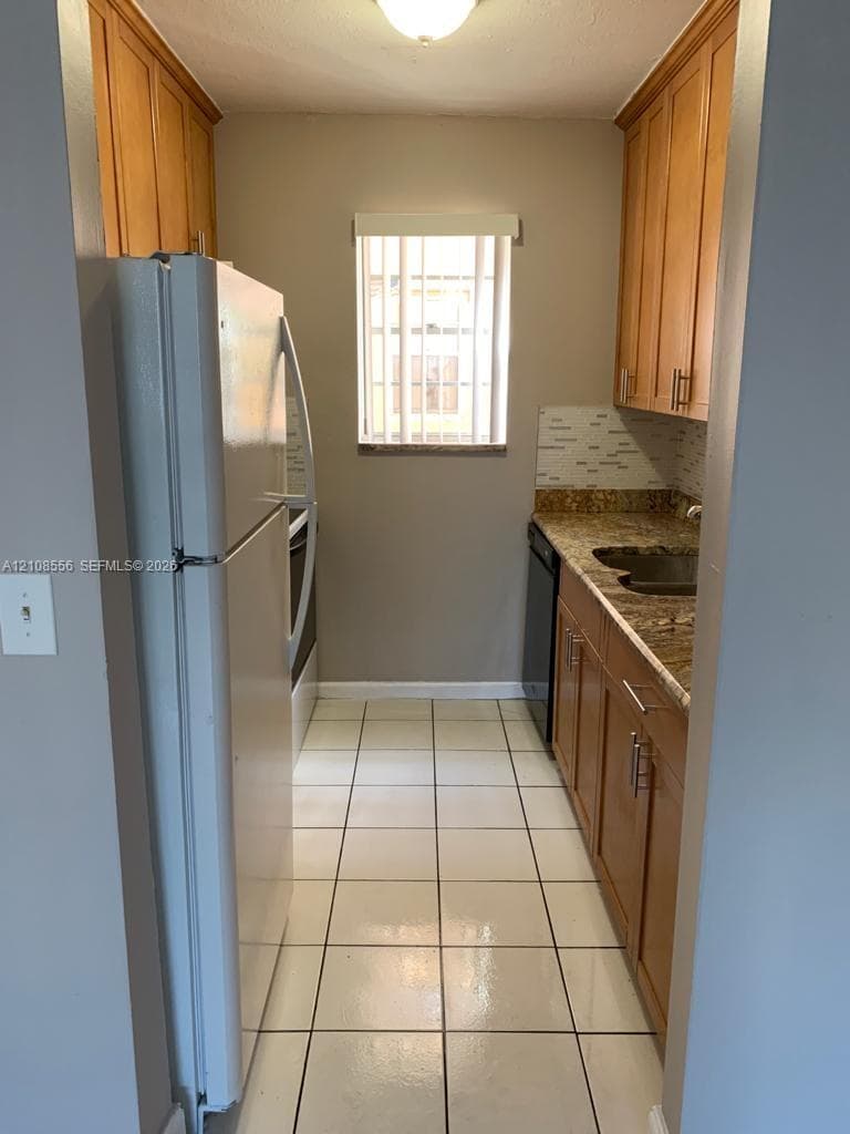 Kitchen featuring natural wood cabinetry, stone countertops, tiled backsplash, large-format tile flooring, and a window