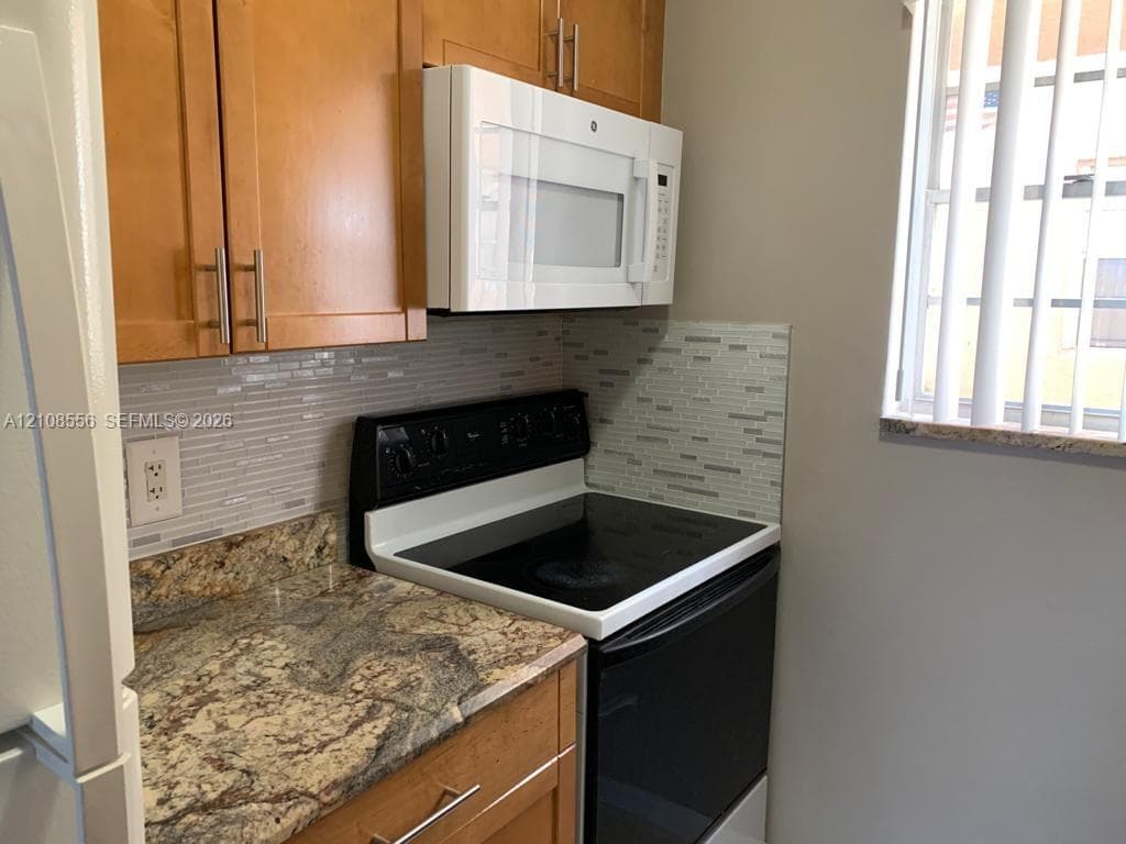 Kitchen featuring wood-finish cabinetry with bar pulls, stone-patterned countertops, a tiled linear mosaic backsplash, and a built-in over-the-range microwave