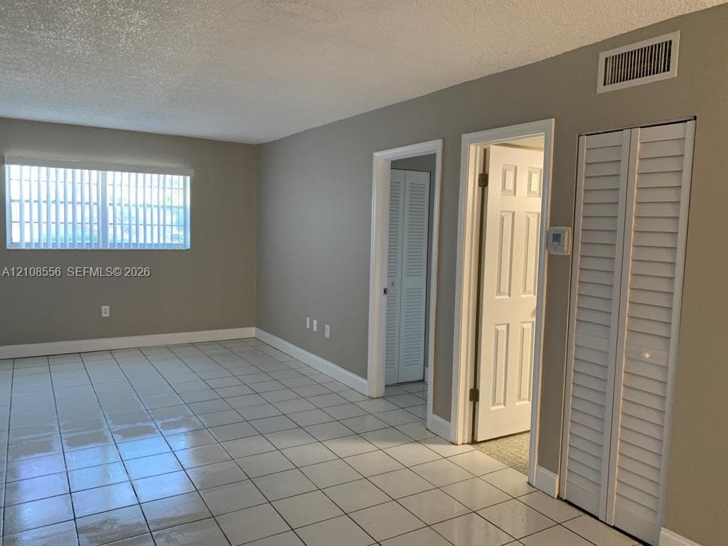 Neutral-toned interior space featuring light-colored ceramic tile flooring, a large window with vertical blinds, and dual white louvered closet doors
