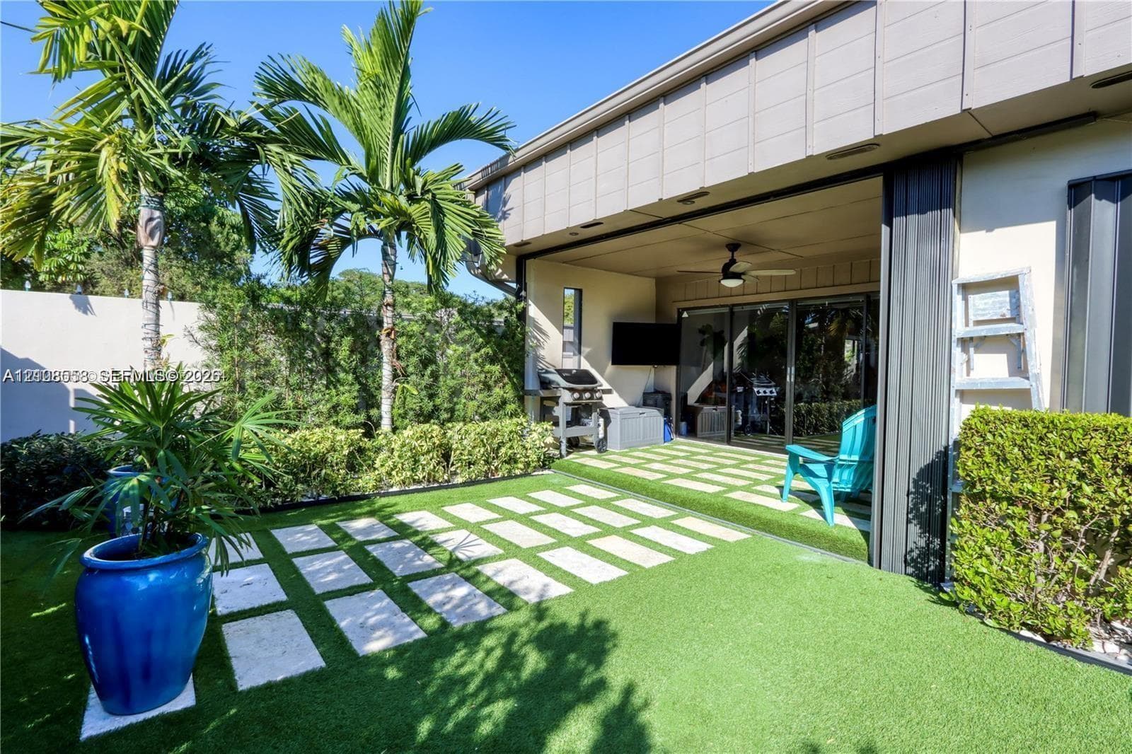 Outdoor patio featuring synthetic turf, light-toned stone pavers, a covered ceiling with an integrated fan, and lush perimeter landscaping