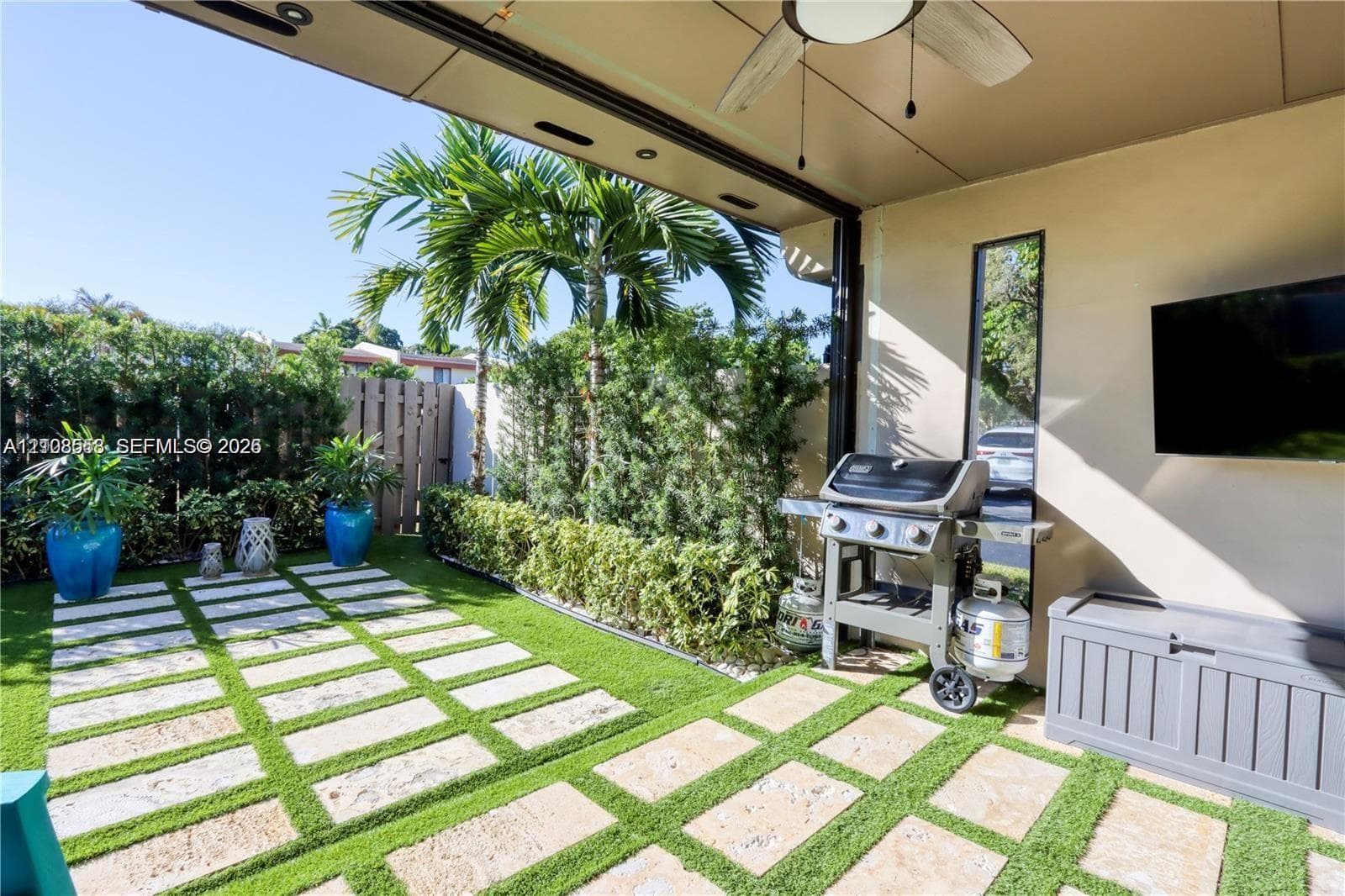 Covered patio with recessed lighting and a ceiling fan, featuring stone pavers with inset synthetic turf, privacy fencing, and perimeter landscaping