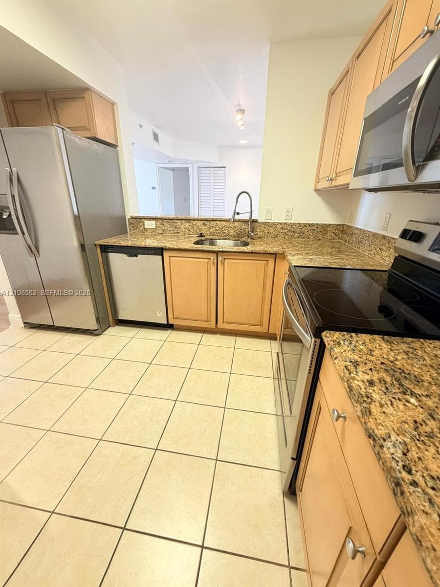 Kitchen featuring natural-finish wood cabinetry, stone-patterned countertops, stainless steel appliances, and tile flooring