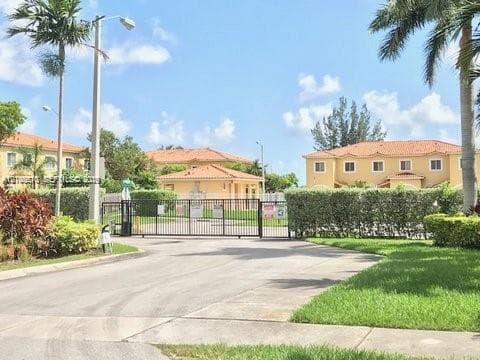 Gated entrance featuring a metal security gate, concrete driveway, and mature perimeter landscaping