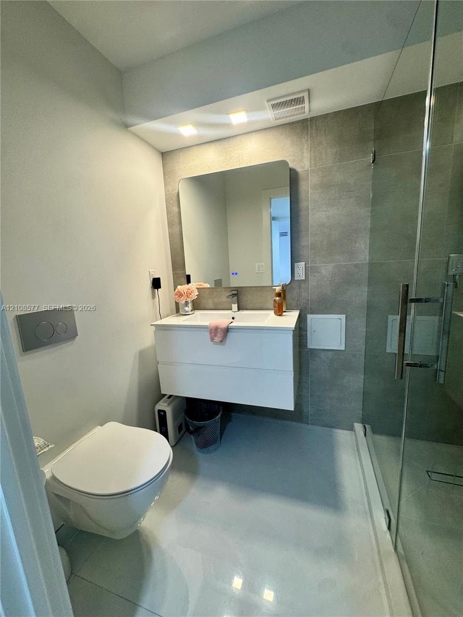 Floating vanity with a white countertop and integrated sink, wall-mounted toilet with a flush plate, and walk-in glass-enclosed shower with large-format grey wall tile