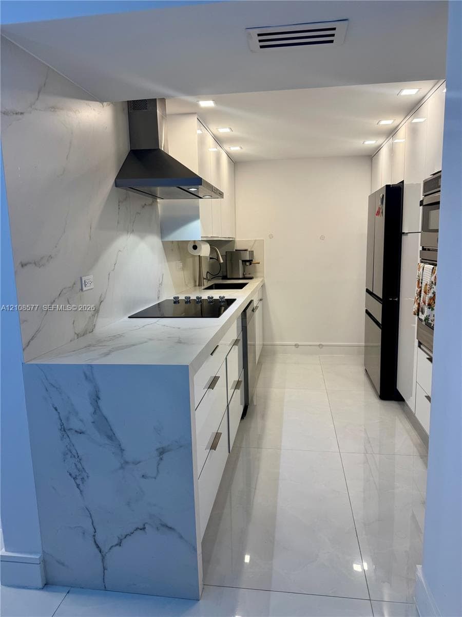 Sleek kitchen featuring high-gloss cabinetry, stone-finish countertops with matching full-height backsplash, polished tile flooring, and integrated stainless steel appliances including a range hood