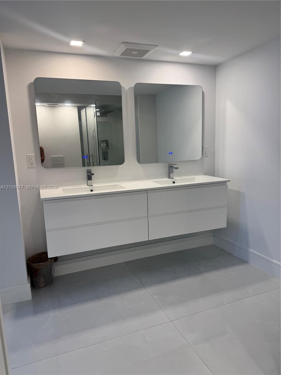 Double floating vanity featuring a white composite countertop, dual integrated sinks, chrome faucets, and two rectangular wall-mounted mirrors with integrated lighting, set against large-format tiled flooring