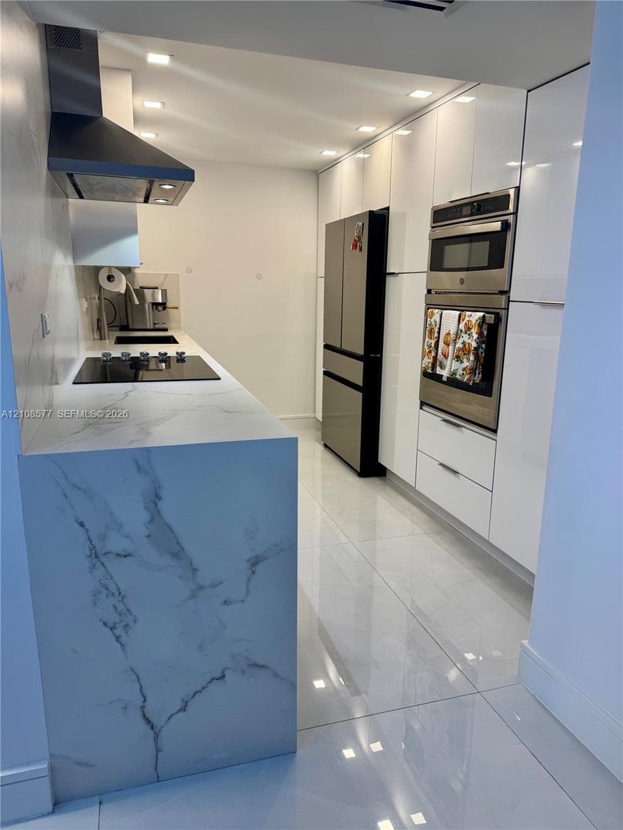 Modern kitchen featuring white flat-panel cabinetry, waterfall-edge stone-finish countertop, stainless steel built-in double oven, glass cooktop, and large-format tile flooring