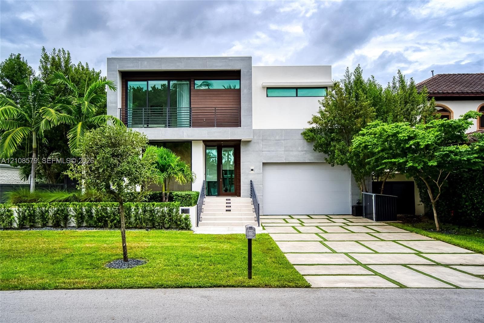 Contemporary multi-level exterior featuring smooth stucco and stone-finish cladding, horizontal wood paneling, metal balcony railing, and a grid-patterned concrete paver driveway